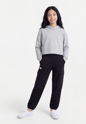 Jeune fille aux longs cheveux foncés portant un sweat à capuche gris court, un pantalon cargo noir avec un logo, des chaussures blanches sans lacets, debout les mains dans les poches.