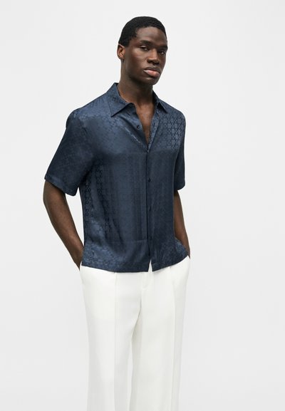 Άνδρας που φοράει σκούρο μπλε πουκάμισο με κοντά μανίκια και διακριτικό μοτίβο, καθώς και λευκό παντελόνι φαρδιάς γραμμής, με τα χέρια στις τσέπες.