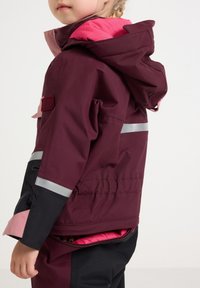 Bordeauxjack met roze accenten, reflecterende strepen en een capuchon. Beschikt over verstelbare manchetten en een ritszak met een gestreepte voering.