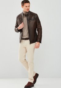 Brown leather jacket with a stand collar, zippered pockets, and textured finish. Worn over a light brown turtleneck and cream-colored pants.