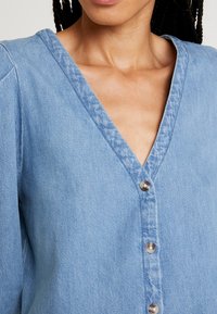 Camicia di jeans blu chiaro con scollatura a V, maniche lunghe e cinque bottoni metallici sul davanti; tessuto strutturato con design casual.
