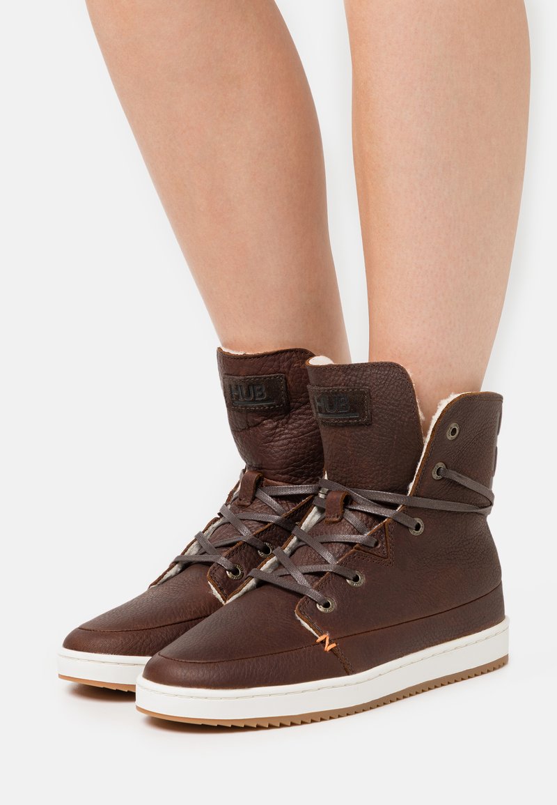 Brown leather high-top sneakers with gray laces, white sole, and fur lining. Features a stitched design and branding on the tongue.