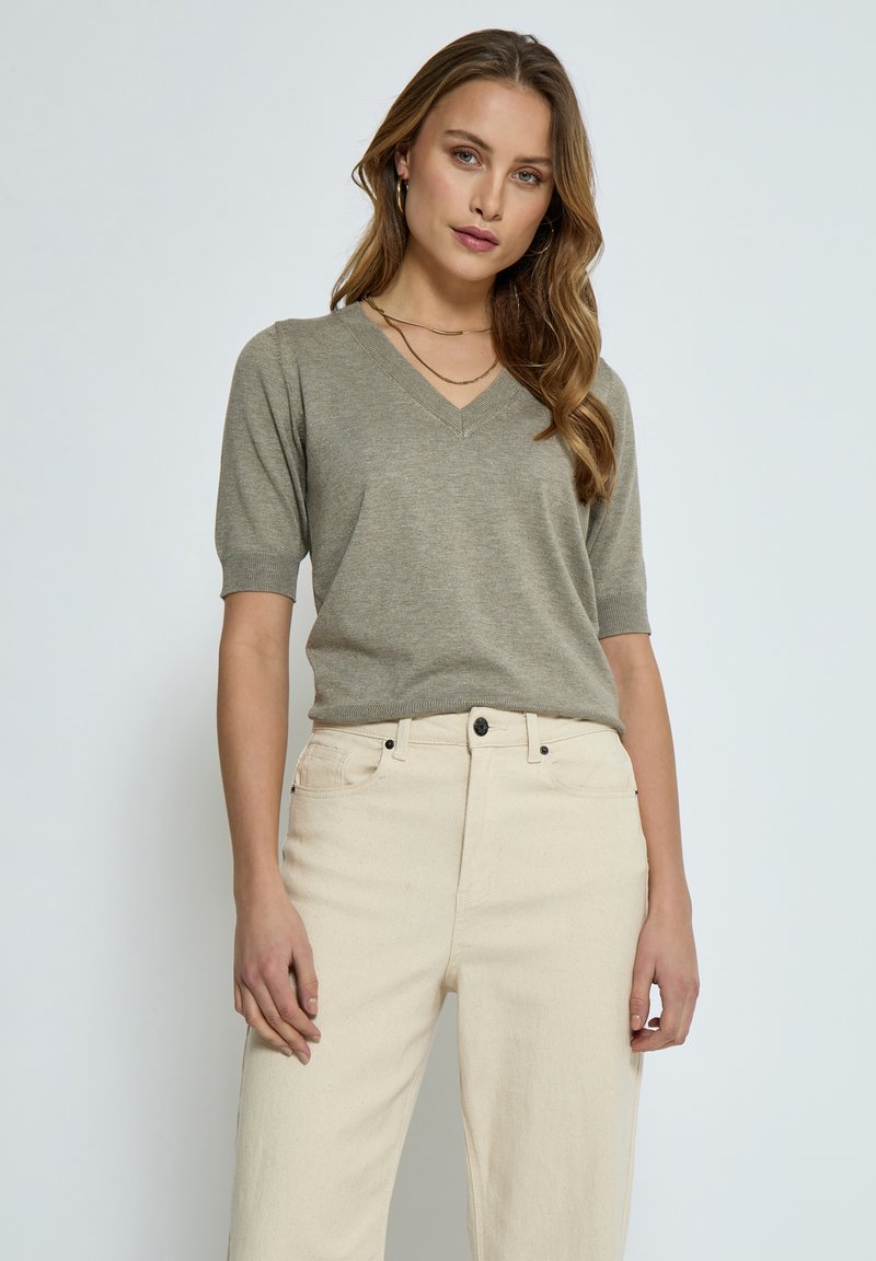 Maglia a V verde oliva in maglia con maniche corte, abbinate a jeans beige chiaro a vita alta. Design semplice, texture morbida, dettagli minimi.