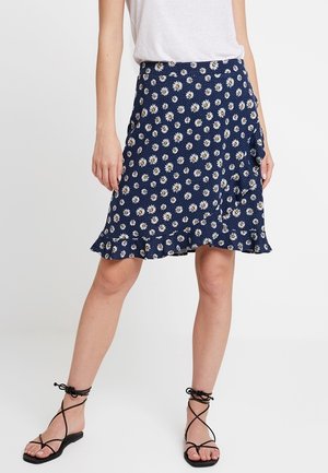 Kobieta w granatowej spódnicy z białym wzorem stokrotek i falbaną na dole, w połączeniu z białym topem i czarnymi sandałami na paski.