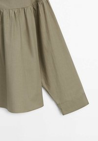 Chemise à manches longues vert olive avec coutures froncées au-dessus du poignet et un poignet boutonné sur un fond blanc uni.