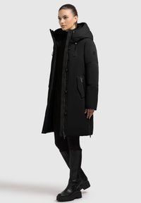Black puffer coat with a hood, featuring a zipper closure, side pockets, and textured fabric. Paired with knee-high black boots.