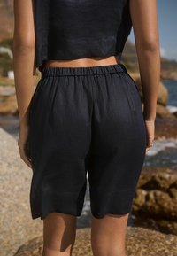 Pantalones cortos de tela oscura y texturizada con cintura elástica, con un ajuste holgado y dobladillo con borde en forma de ondas, vistos desde atrás con un fondo rocoso.
