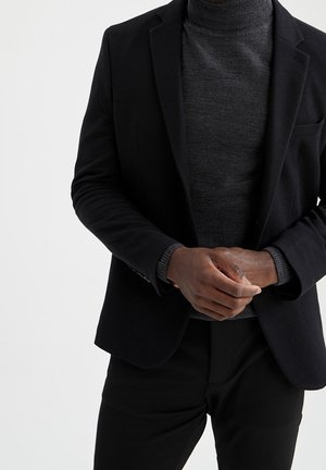 Mann trägt schwarzen Blazer und dunkelgrauen Rollkragenpullover, die Hände vor dem Körper verschränkt.