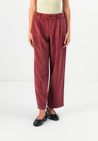 I pantaloni a palazzo in corduroy rosso presentano un alto girovita, pieghe frontali e una chiusura con bottone, abbinati a scarpe basse nere.