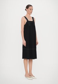Rochie neagră fără mâneci, cu bretele late, având cusături în contrast, formă A și tiv până la genunchi. Asortată cu pantofi mule albi cu vârf ascuțit.
