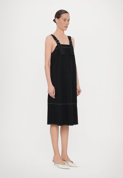 Crna haljina bez rukava sa širokim trakama, s kontrastnim šavovima, A-linijski oblik i dužina do koljena. Uparena s bijelim cipelama na špic.