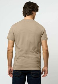 Vyras su tamsiais plaukais, dėvintis paprastą smėlio spalvos trumpomis rankovėmis marškinėlį ir tamsiai mėlynas džinsines kelnes, matomas iš nugaros baltame fone.