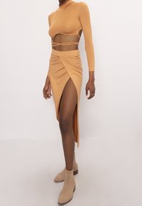 Top ajustado de manga larga en color beige con detalles de corte en los laterales, combinado con una falda maxi drapeada a juego que presenta una abertura alta en el muslo. Botines neutros hasta el tobillo.