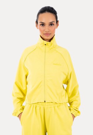 Frau trägt eine leuchtend gelbe Reißverschlussjacke und passende Hose, steht mit den Händen in den Taschen vor einfachem Hintergrund.