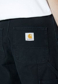 Černé plátěné kalhoty s velkou zadní kapsou, na které je čtvercová bílá etiketa s žlutým logem. Po obvodu kapsy přecházejí našité detaily.