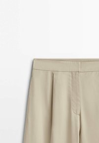 Pantalon beige taille haute avec des plis sur le devant et une ceinture cousue, présenté sur un fond blanc.