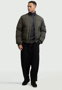 Άνδρας που φοράει ελαιόχρωμο χνουδωτό μπουφάν, σκούρο γκρι πουλόβερ με πλέκτο φουλάρι, χαλαρούς μαύρους παντελόνες και μαύρα παπούτσια, στέκεται με τα χέρια στις τσέπες.