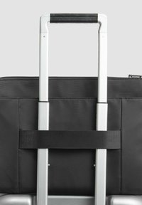 Valigia in tessuto nero con manico telescopico in alluminio, dotata di impugnatura testurizzata e cinghia di sicurezza. Base argentata visibile nella parte inferiore.