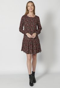 Blumengemustertes Kleid mit langen Ärmeln, rundem Ausschnitt, schwingender Taille und gerafftem Saum. Die Farben umfassen Rot, Schwarz und Grün. Getragen mit schwarzen Knöchelstiefeln.