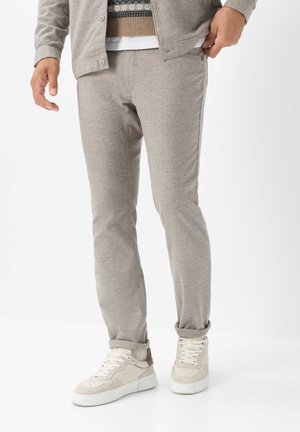 Mann trägt hellgraue Freizeithose, beige und weiße Sneakers und ein teilweise sichtbares Lagenoutfit, stehend vor weißem Hintergrund.