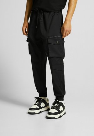 Mann trägt schwarze Cargo-Joggerhosen mit mehreren Taschen und schwarz-weißen Sneakers, steht vor einem hellgrauen Hintergrund.
