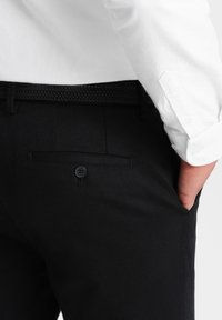 Pantaloni neri in tessuto texture, con una tasca posteriore con chiusura a bottone e una cintura nera intrecciata. Camicia bianca parzialmente visibile.