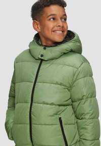 Grüne gepolsterte Jacke mit abnehmbarem Kapuzen, ausgestattet mit einem Frontreißverschluss und zwei Seitentaschen. Das Innere hat ein schwarzes Logoband.