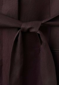 Ciemnobrązowa tkanina o gładkiej fakturze z paskiem wiązanym w talii, podkreślająca czysty, uporządkowany design oraz ukryte boczne kieszenie.