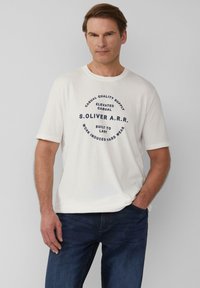 Witte katoenen T-shirt met korte mouwen, voorzien van een cirkelvormig marineblauw grafisch tekstontwerp in het midden. Gedragen met marineblauwe spijkerbroek.