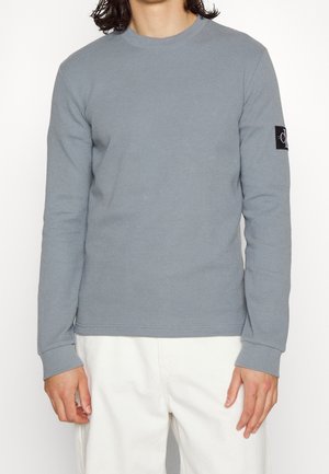 Mężczyzna ubrany w jasnoszarą, teksturowaną, długą koszulę z czarną naszywką z logo na lewym rękawie i białe spodnie, stojący na białym tle.