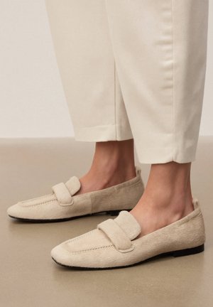Füße tragen beigefarbene Wildleder-Loafers, kombiniert mit cremefarbenen Cropped-Hosen vor neutralem Boden- und Wandhintergrund.