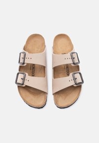 Μπεζ σανδάλια Birkenstock με δύο ρυθμιζόμενες μαύρες αγκράφες και διαμορφωμένες φελούς σόλες σε λευκό φόντο.