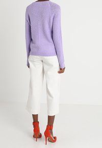 Pull en maille lavande avec texture côtelée, pantalon blanc raccourci, et sandales à talons hauts rouge vif avec brides à la cheville et orteils ouverts.
