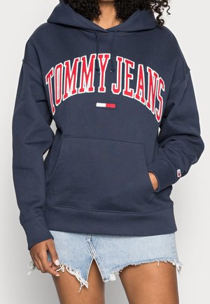 Osoba w granatowej bluzie z kapturem z "TOMMY JEANS" czerwonymi i białymi literami oraz w jasnoniebieskiej postrzępionej dżinsowej spódnicy.