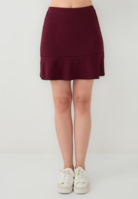 Femme portant une jupe courte violet foncé et des baskets blanches, se tenant devant un fond léger et uni.