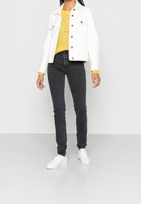 Frau in einer weißen Jeansjacke, einem gelben Strickpullover, dunkelgrauen Skinny-Jeans und weißen Sneakers, die auf einem grauen Boden steht.