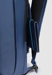 Zaino blu con trama, dotato di una cerniera nera, con un portafoglio scuro sottile parzialmente visibile in una tasca laterale. Spallacci regolabili.