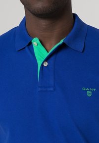 Blå pikétröja med grön krage, två knappar och en liten grön "GANT"-logotyp på bröstet. Tillverkad av texturerad bomullstyg.