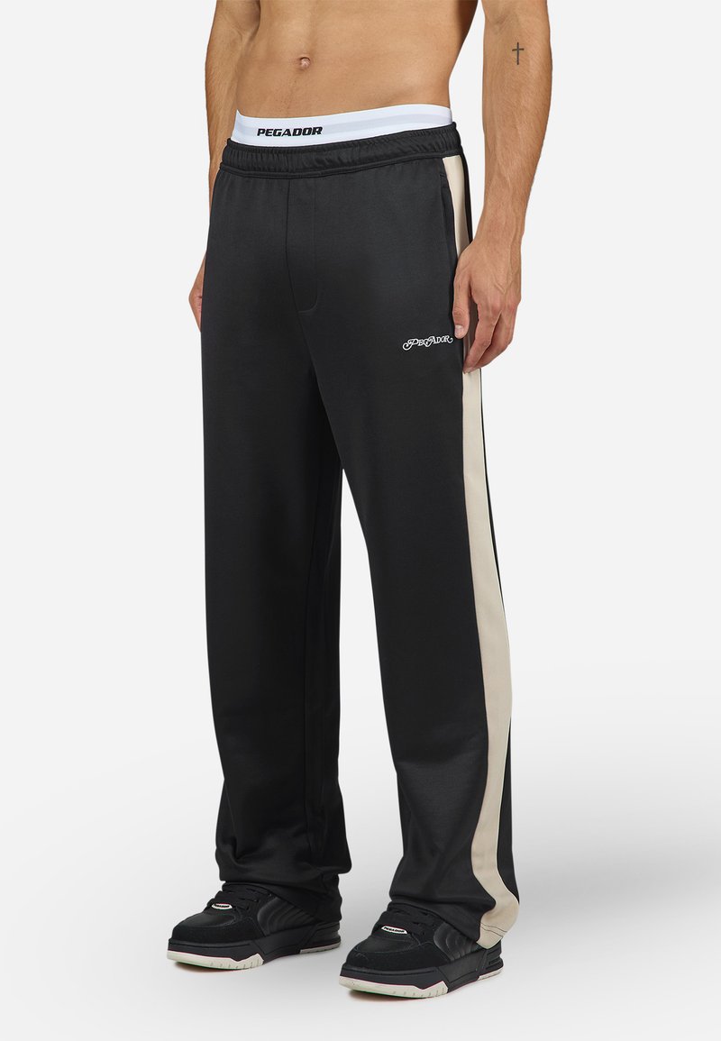 Sorte træningsbukser med beige sidelogo-striber, elastisk talje med logo, glat stof og to sidelommer. Båret med sorte sko.