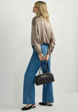 Kobieta stojąca w luźnej bluzce w kolorze taupe, szerokich niebieskich dżinsach, czarnych balerinach na rzemyku, trzymająca czarną torebkę za plecami.