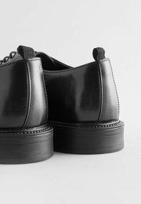 Chaussures en cuir noir avec une finition brillante, bout rond et semelle en caoutchouc épaisse ; présentent des détails cousus et des boucles en tissu au niveau des talons.