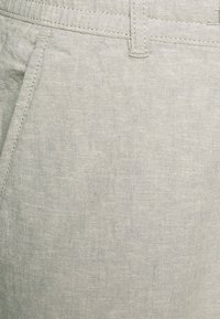 Pantalon en lin gris clair avec une texture lisse, présentant des coutures surpiquées et des poches latérales pour plus de fonctionnalité.