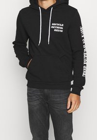 Sudadera negra con cordones blancos y el texto impreso "RECICLAR, REPENSAR, REUTILIZAR" en la parte delantera; mangas largas con "EL FUTURO ERES TÚ" en un lado.