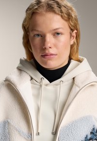 Tânără cu păr blond purtând o jachetă crem din sherpa peste un hanorac crem cu șnururi, pe un fundal neutru.