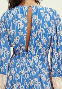 Modré šaty s potiskem palmových stromů v světle růžové barvě, s hlubokým výřezem na zádech a knoflíkovým zapínáním. Tkanina vypadá lehká a texturovaná.