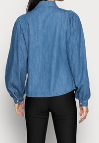 Mujer vestida con una blusa de mezclilla azul de manga larga con mangas abullonadas y pantalones negros ajustados, mostrada de espaldas sobre un fondo simple.