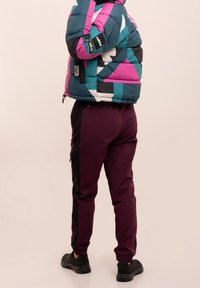 Kleurige pufferjack in teal, roze en zwart geometrisch patroon; gecombineerd met maroon taps toelopende broek met zwarte zijkanten en zwarte schoenen.