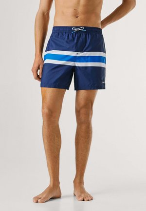 Uomo che indossa pantaloncini da bagno blu navy con strisce orizzontali bianche e blu, in piedi a piedi nudi su uno sfondo semplice.