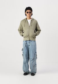 Olivgrüne Zip-Up- Hoodie mit zwei Vorderaschen, kombiniert mit lockeren blauen Denim-Cargohosen, die über mehrere Taschen und eine lässige Passform verfügen. Schwarze Schuhe.
