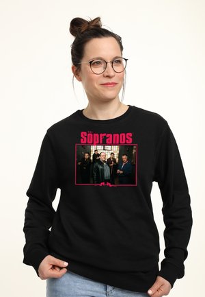 Kvinde med briller iført en sort sweatshirt med et billede af The Sopranos-castet og logoet, kigger til siden med et let smil.